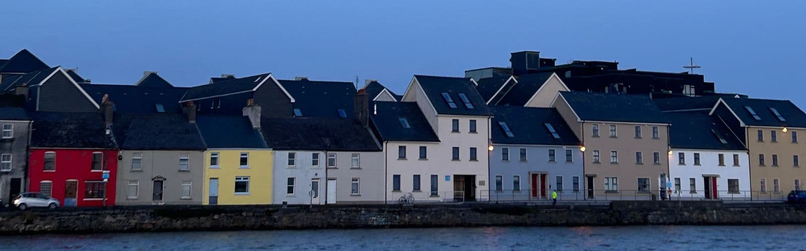 A row of brightly colored houses sits on the edge of a harbor at dusk. 